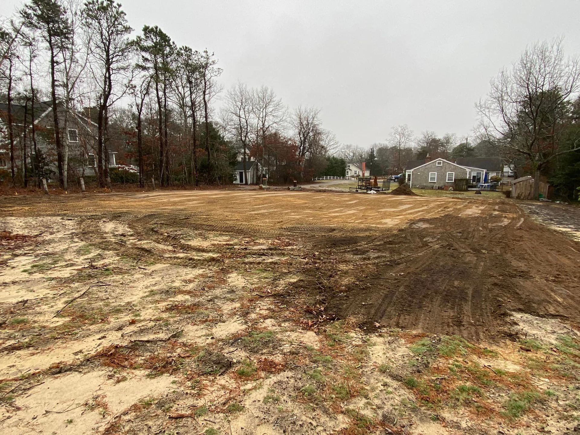 Cleared dirt lot, prepared for construction, with trees and houses in the background on a cloudy day.