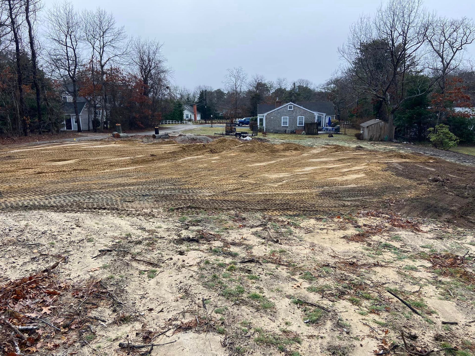 Cleared sandy lot with visible tire tracks, trees in the background, and a small house. Overcast sky.