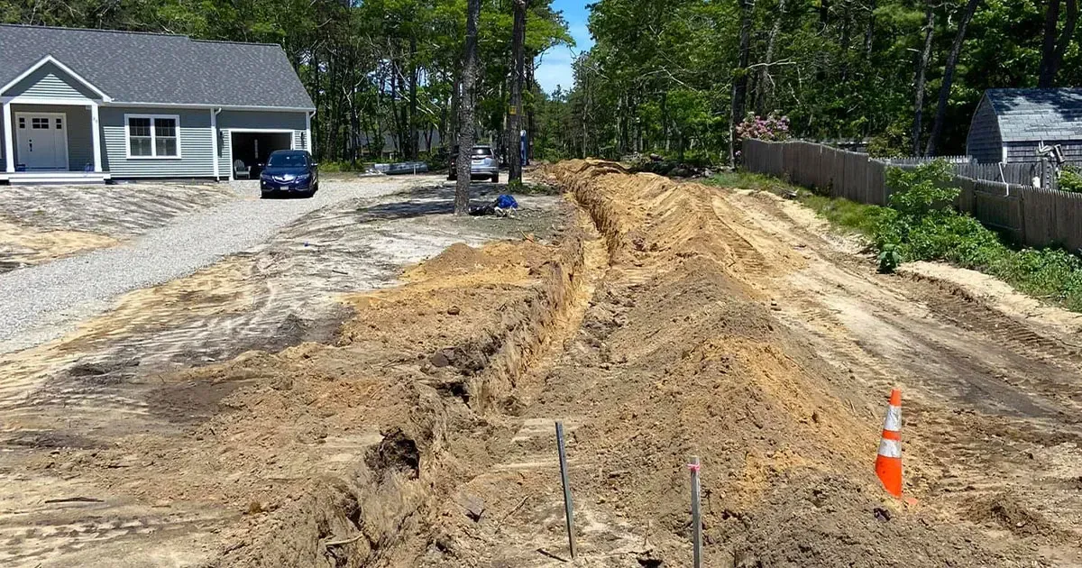 Construction site with trench in the foreground, a house and car in the background, and trees lining the edges.