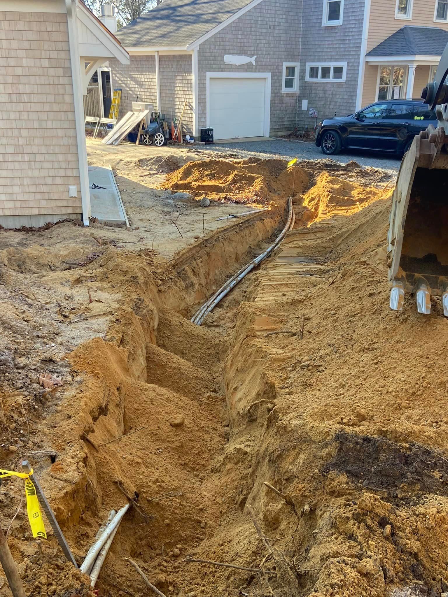Trench dug in dirt with electrical conduit, construction equipment visible. Buildings and a car in background.