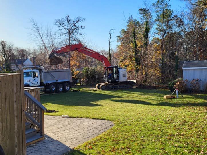 An excavator loading a dump truck with dirt in a grassy yard, near trees and a small shed.