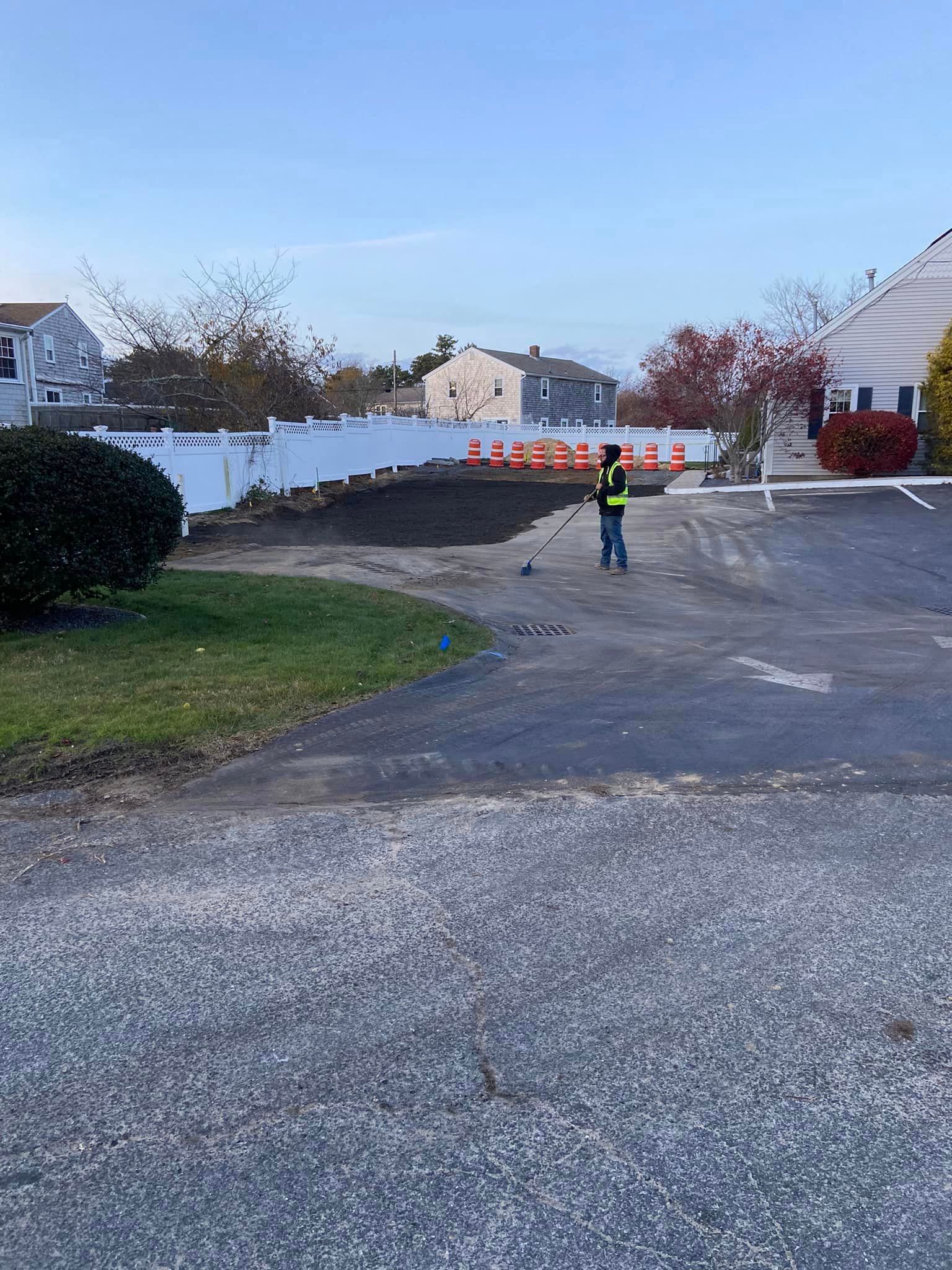 Person raking dirt in a yard near a driveway, with white fence, houses, and construction cones visible in the background.