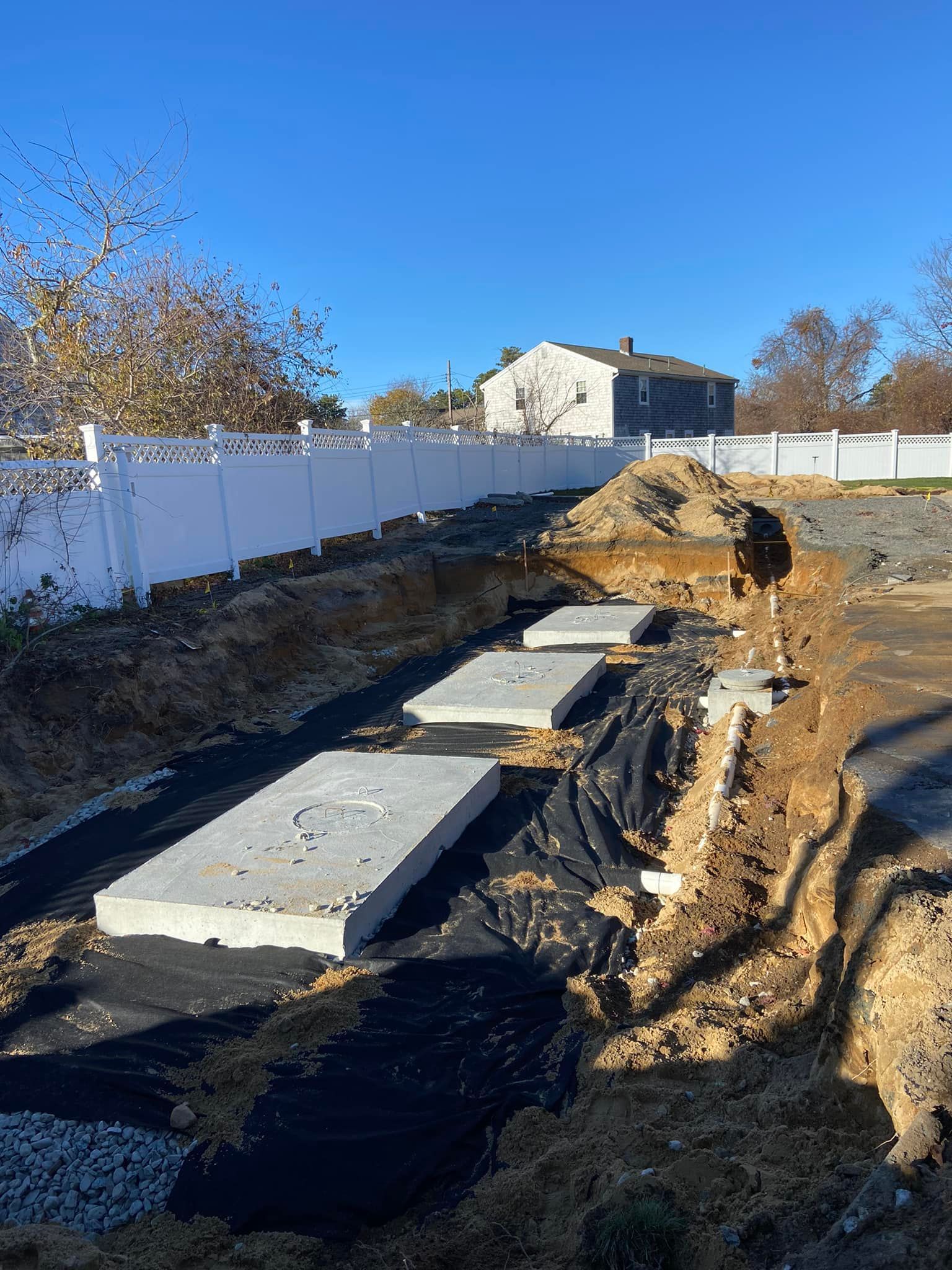 Three concrete septic tank lids in a trench lined with black fabric; a house and white fence in the background.