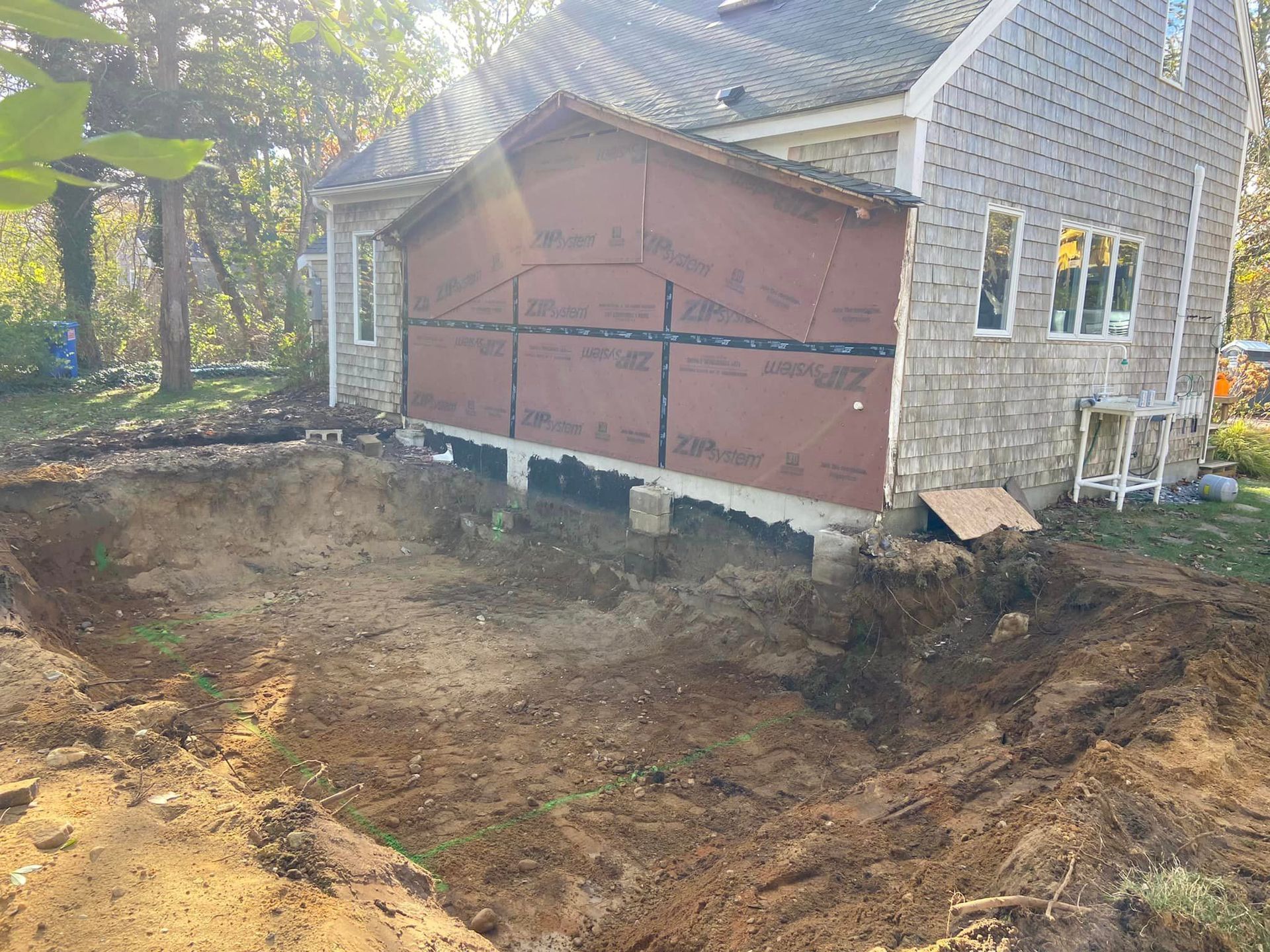 House exterior under construction; excavation in foreground, exposed brown siding, gray shingles, and windows.