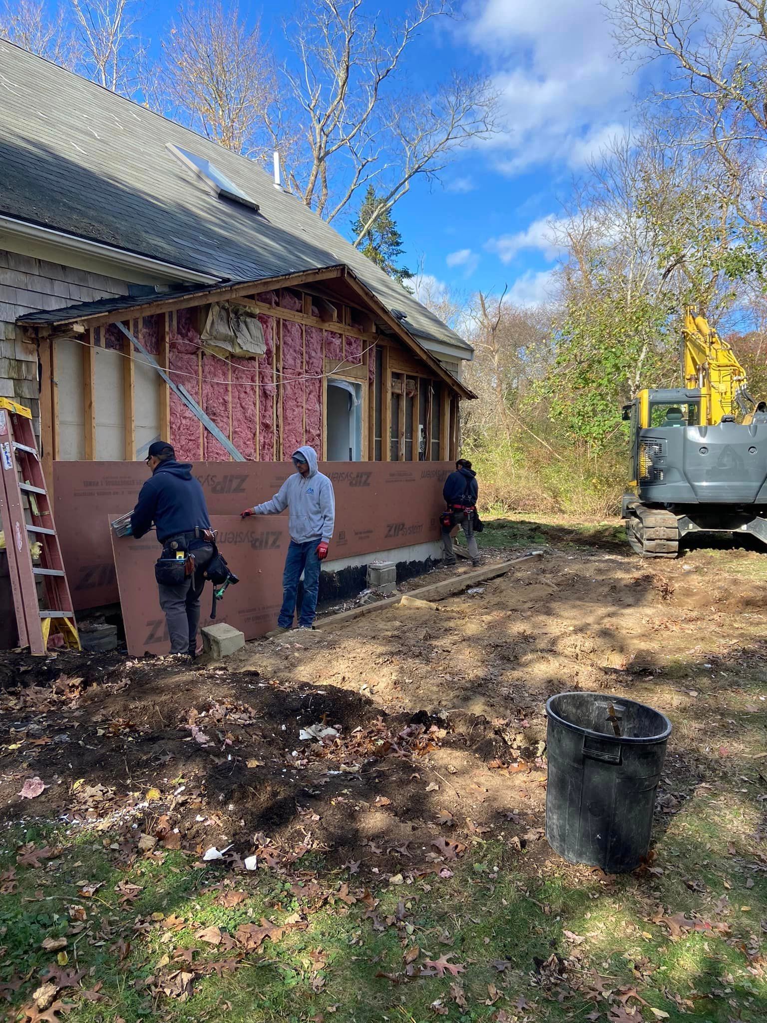 Construction workers installing siding on a house exterior. Excavator, ladder, insulation visible.