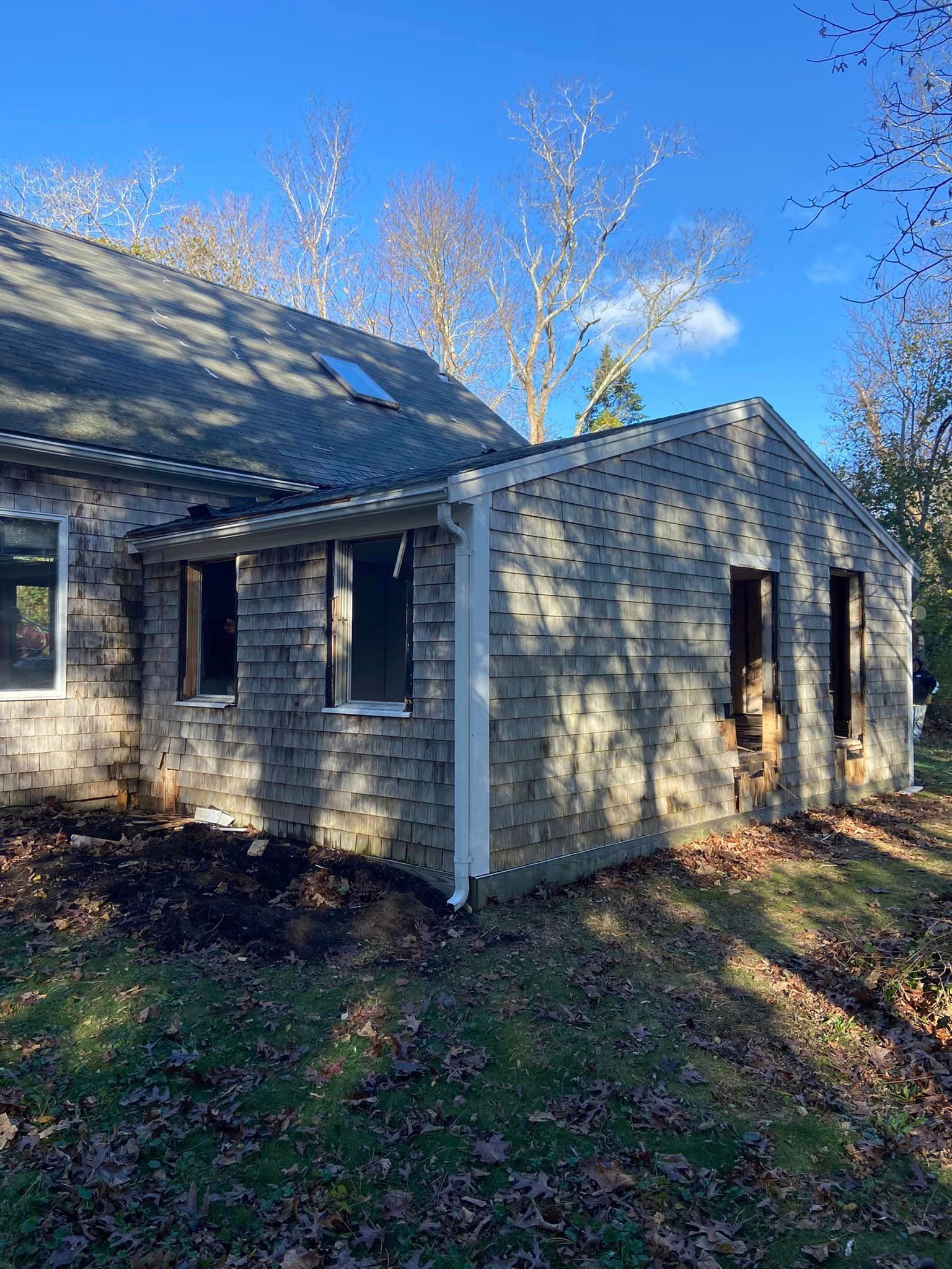 Exterior view of a weathered wooden building with dark roofing and empty window frames, on a sunny day.