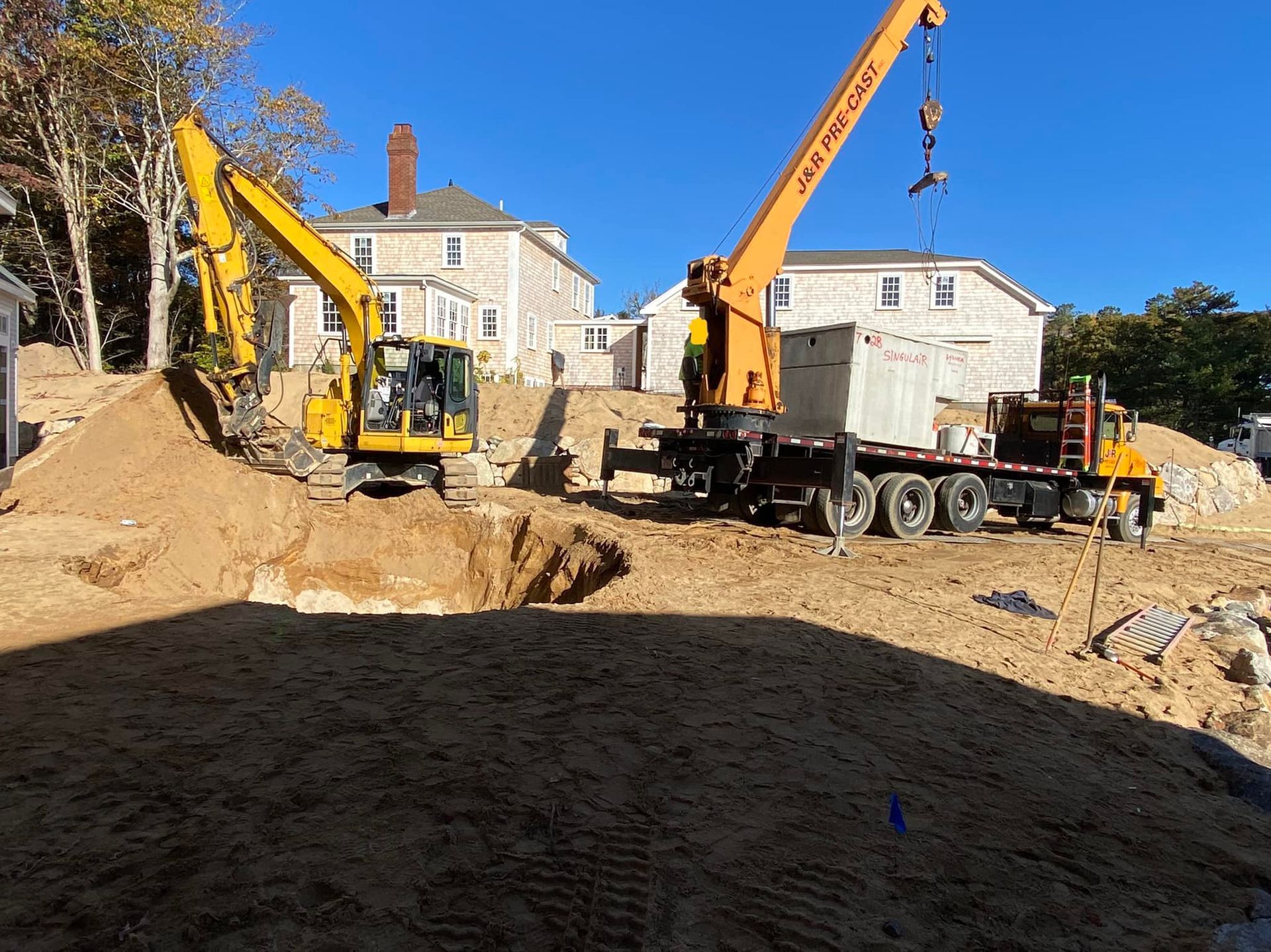 Excavator and crane at a construction site, moving concrete blocks. Sunny day, beige soil, houses in the background.