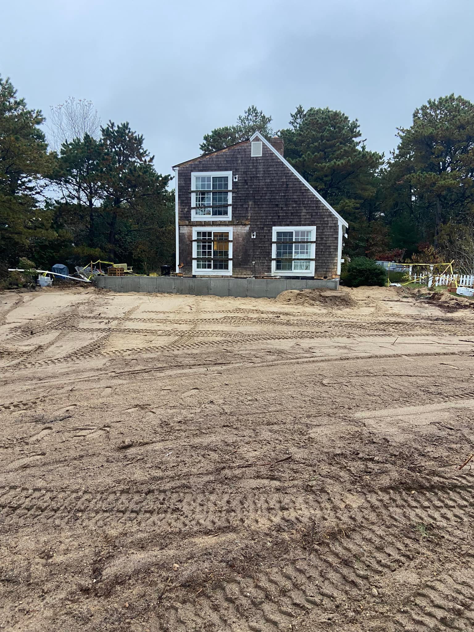 Two-story house with weathered wood siding and large windows, surrounded by trees and a dirt lot.