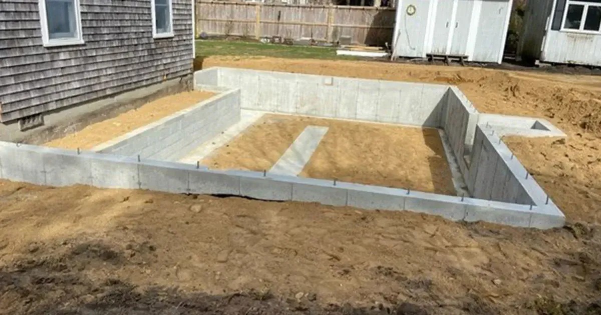 Exterior view of a newly constructed concrete foundation, surrounded by dirt and an existing house.