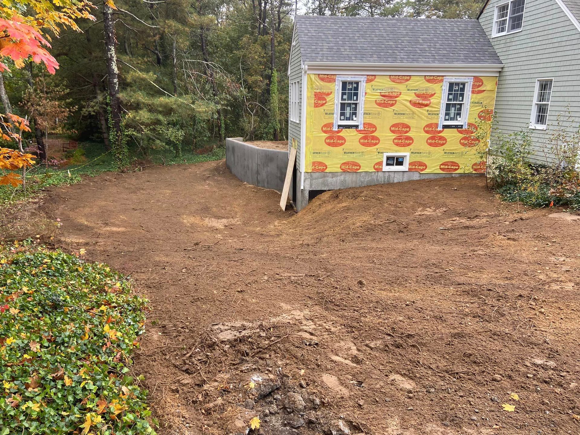 A house with unfinished siding sits next to a dirt yard and a retaining wall, surrounded by trees.