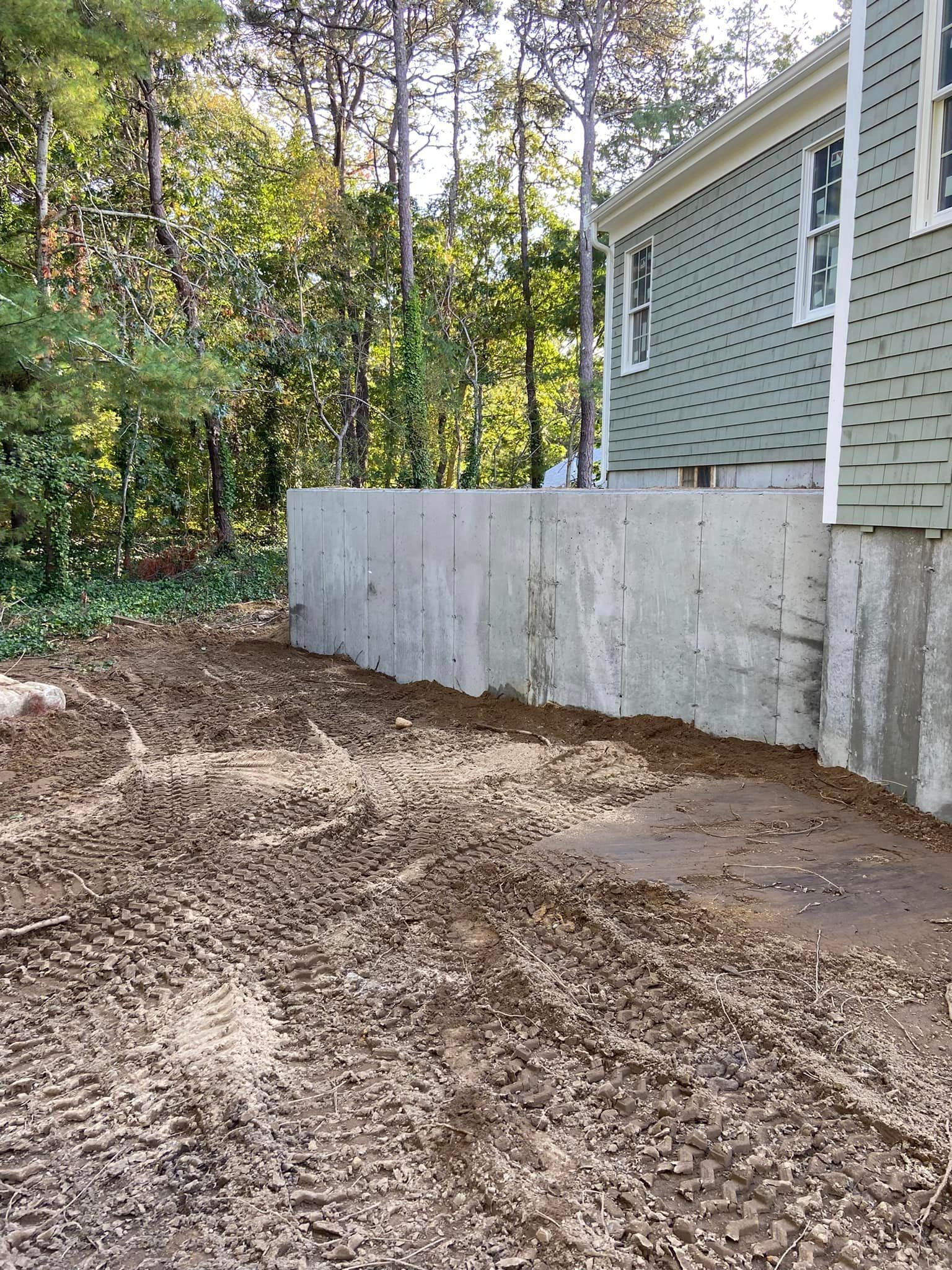 Dirt and gravel area next to a concrete wall and a house with green siding, trees in the background.