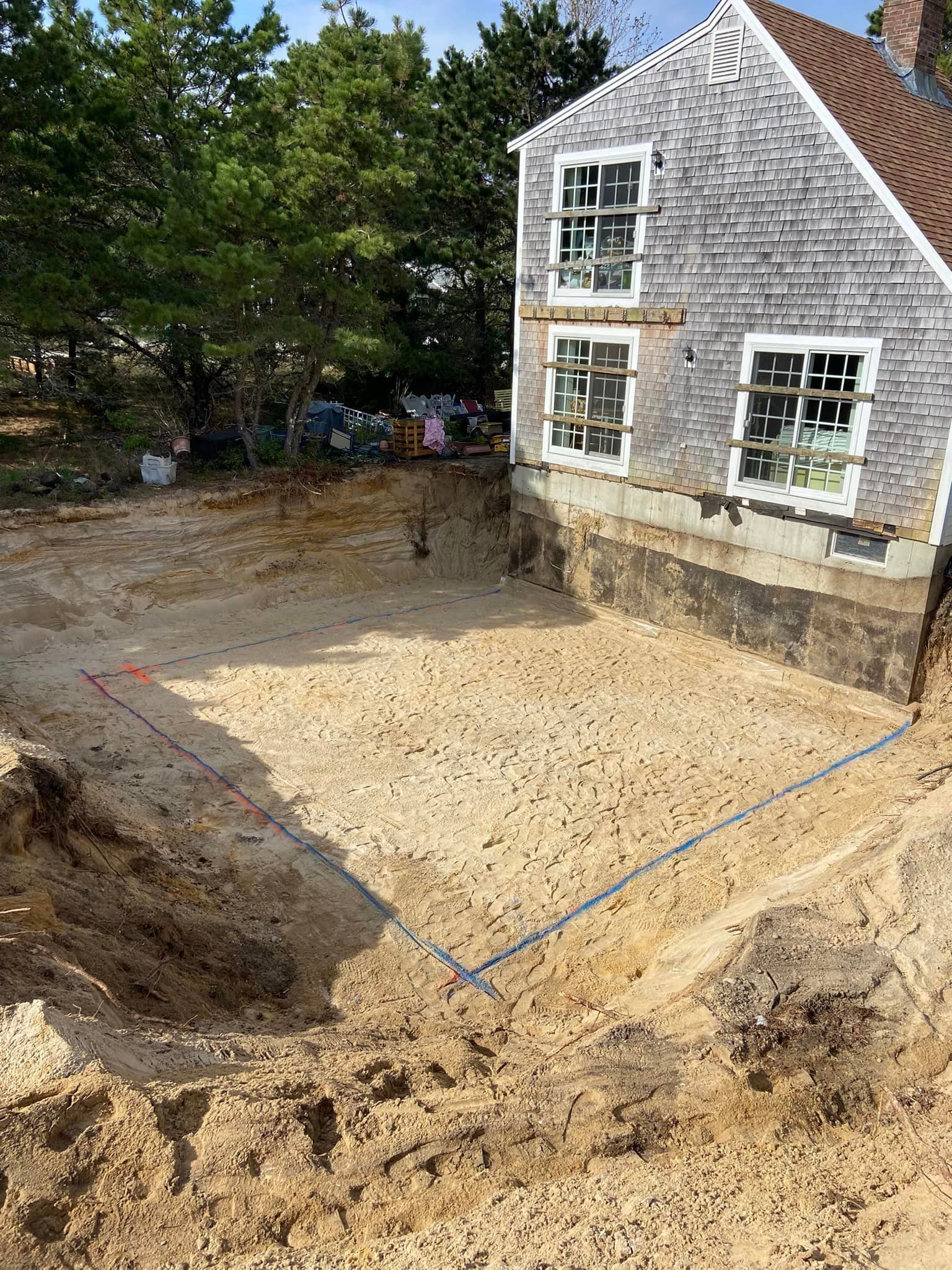 Construction site: excavated rectangular foundation next to a shingled house.