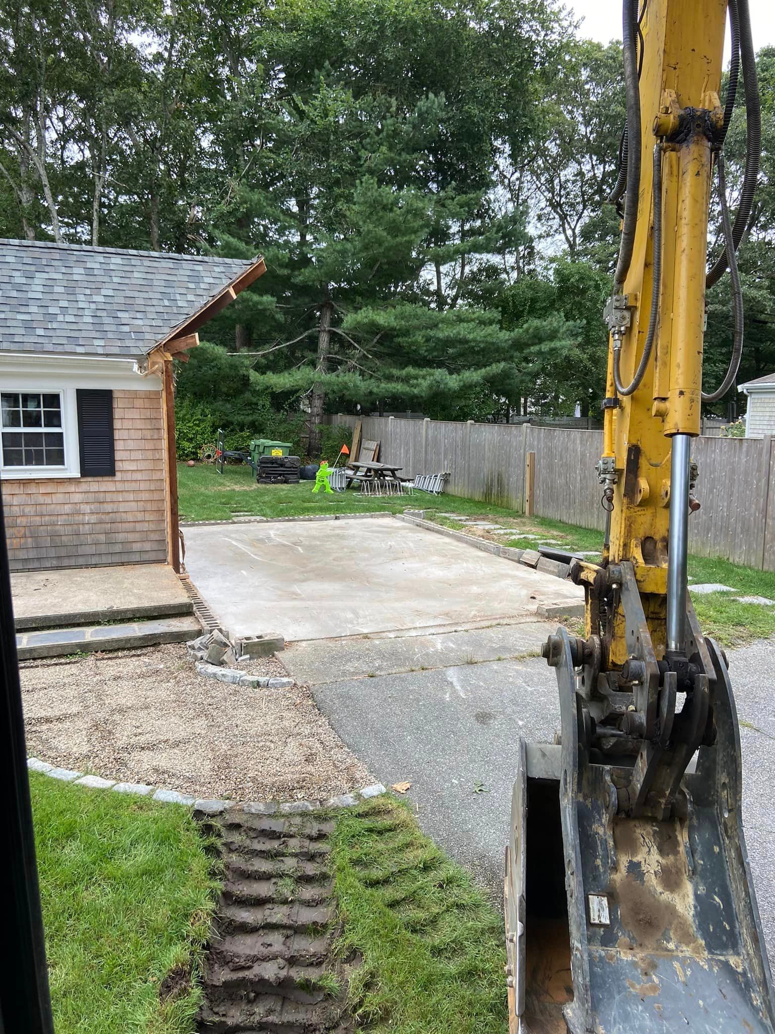 Excavator on a residential lot, digging up a concrete driveway. House and trees are visible in the background.