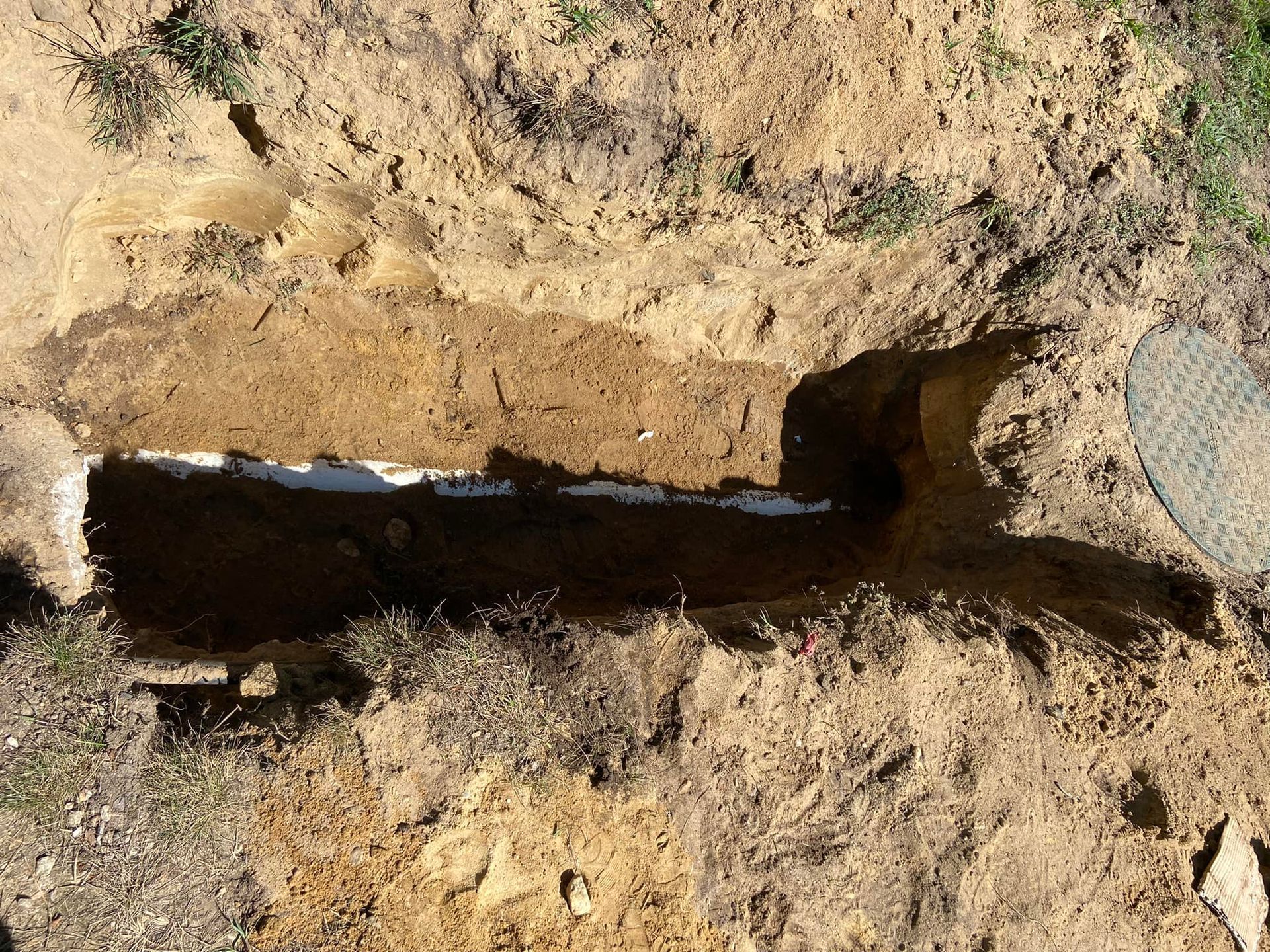 Trench dug in the ground, revealing white pipe; lid of a drain visible on the right.