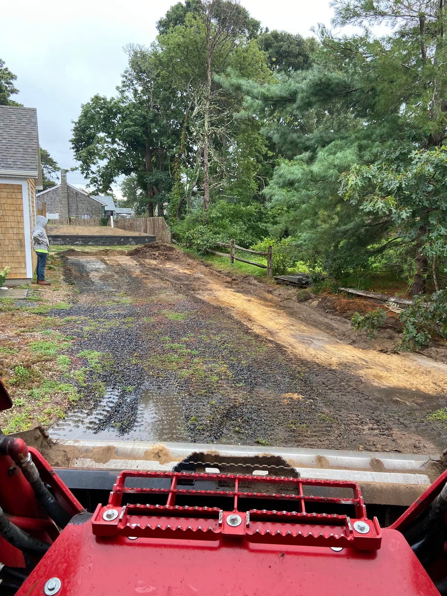 View from a tractor cab of a muddy driveway leading uphill to a house and trees.