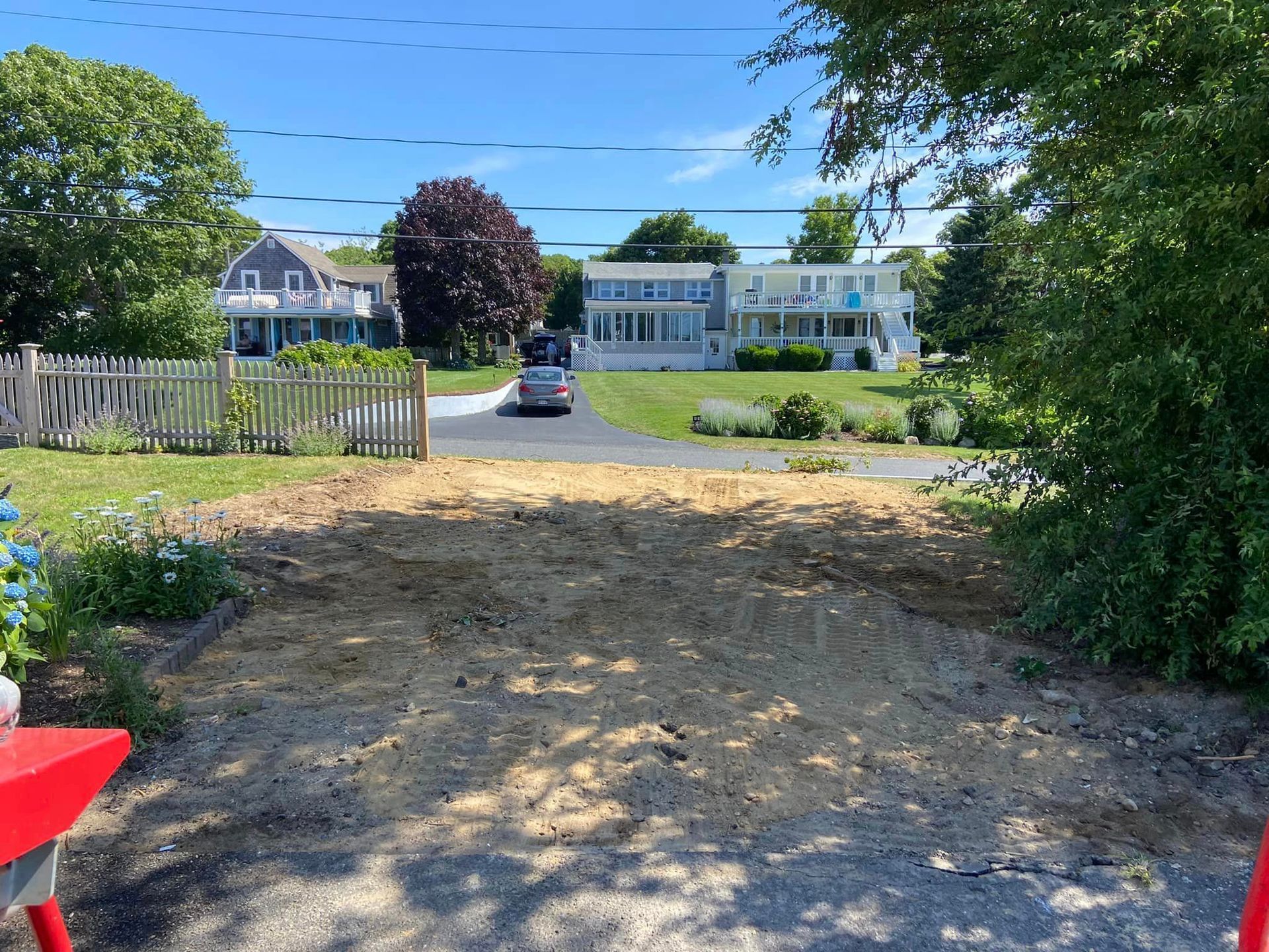Dirt mound in front of driveway, houses and trees in background under blue sky.