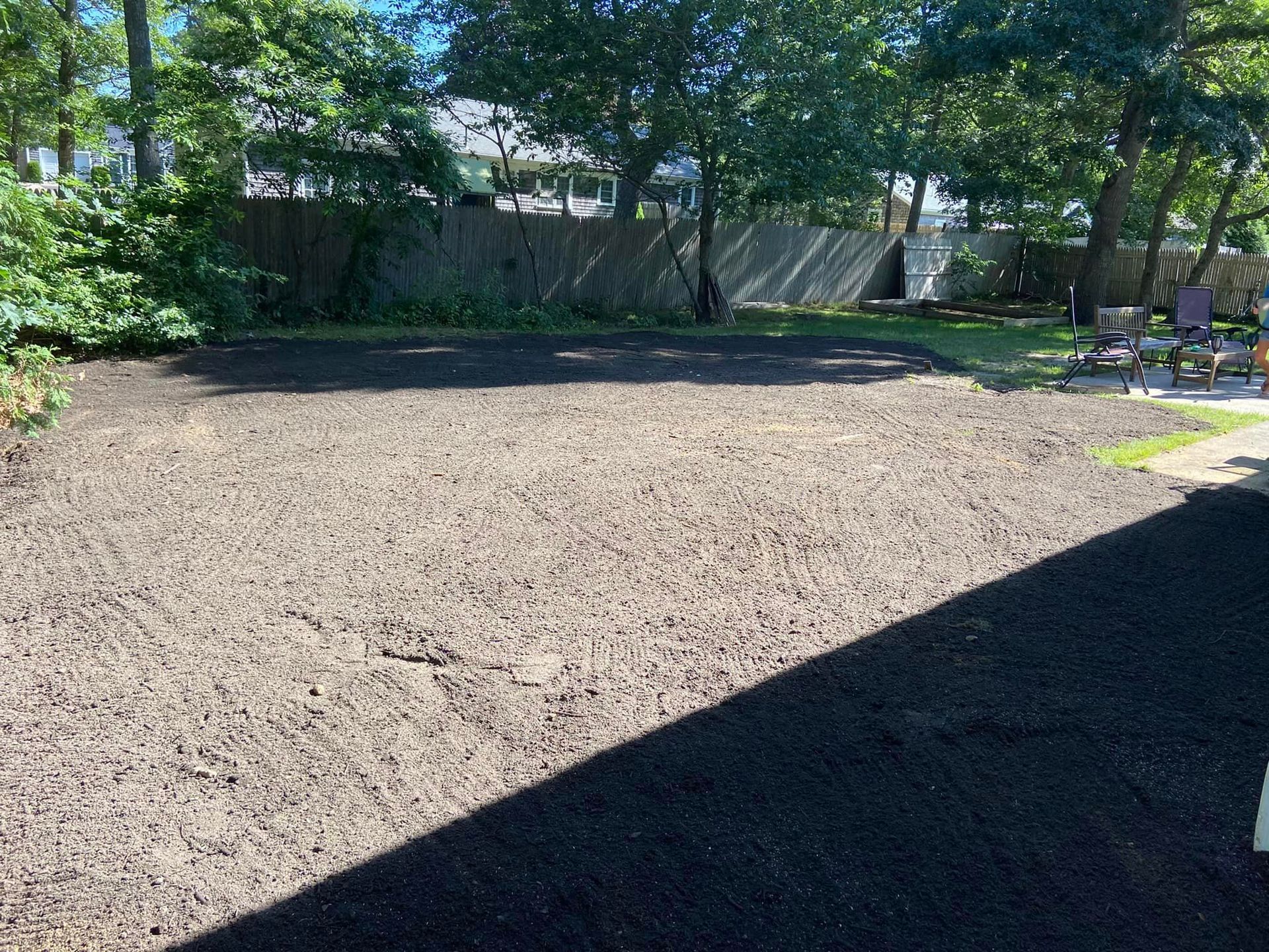 A backyard area covered in dark mulch, with trees and a fence in the background on a sunny day.