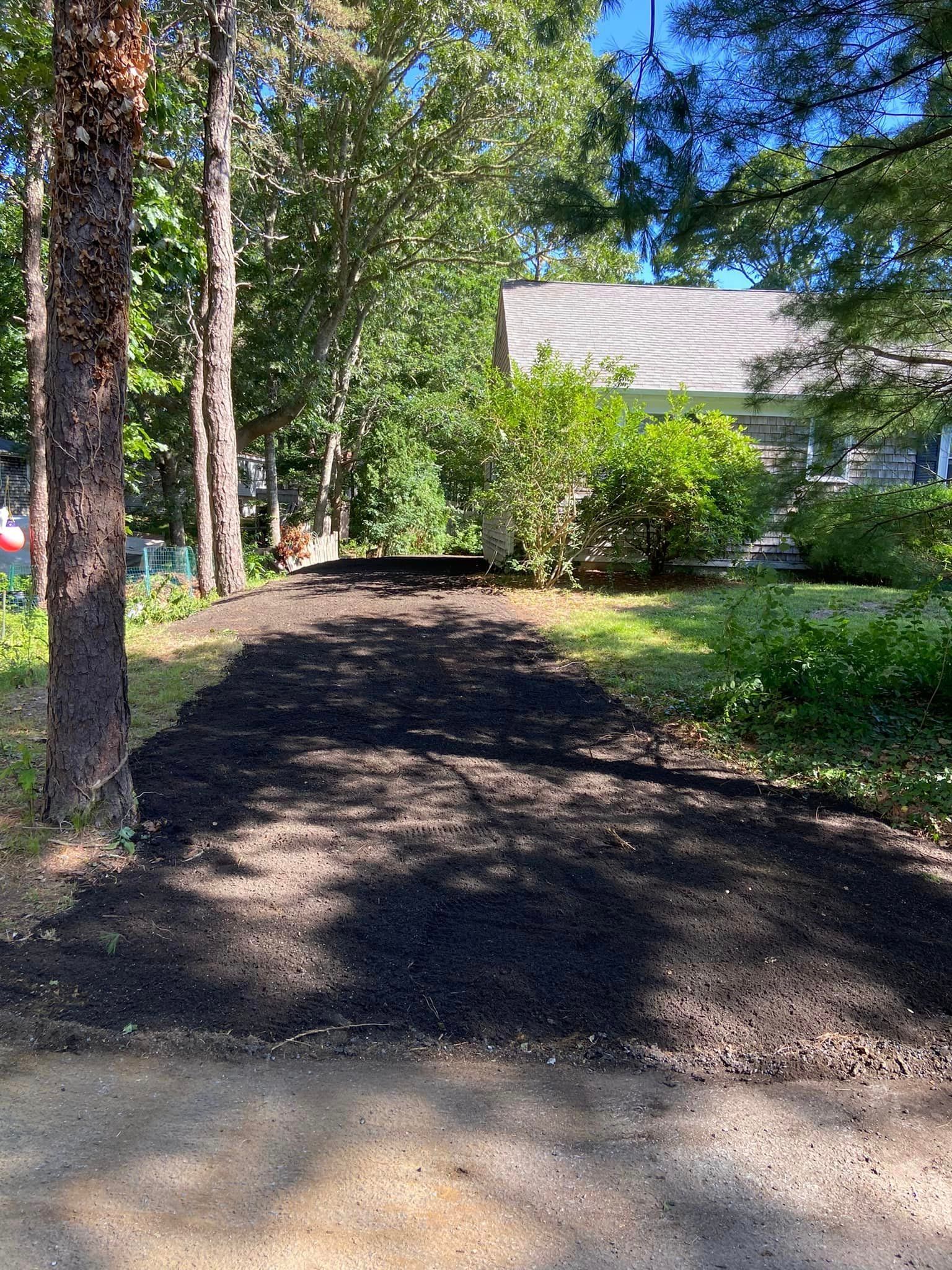 Newly paved asphalt driveway through a wooded area, leading towards a house.