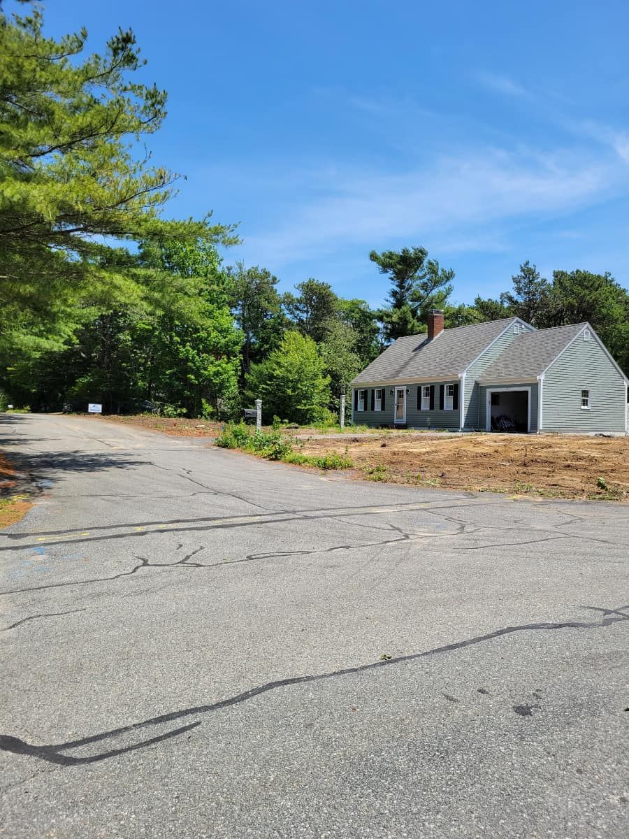 A house on a dirt lot, partially obscured by trees, on a sunny day. Gray roof and siding. Paved road.