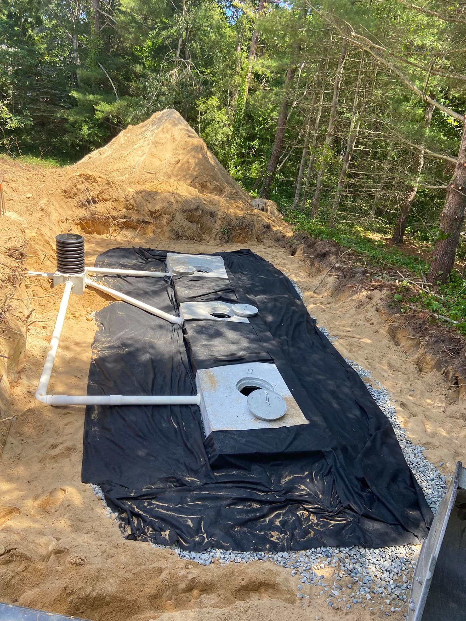 Septic system components in a trench, including tanks and pipes, surrounded by sand and landscaping fabric.