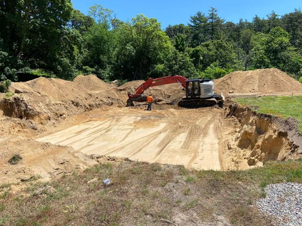 An excavator digging in a large dirt pit; a person stands nearby on a sunny day.