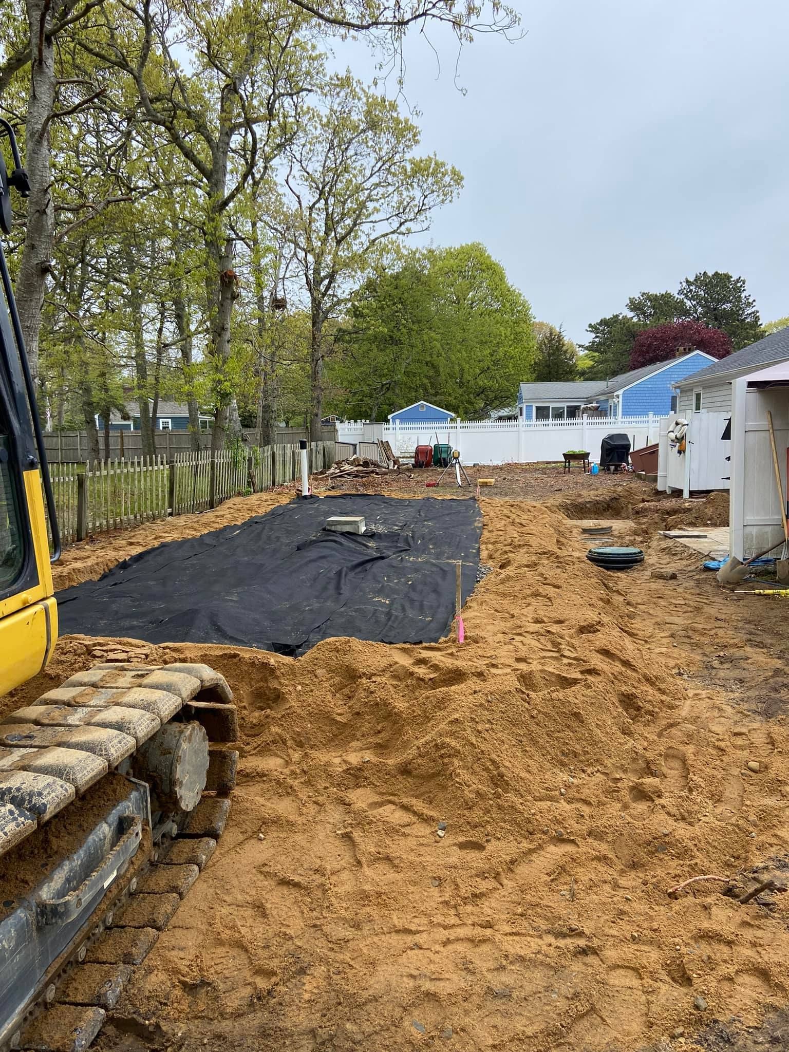 Construction site, excavator beside a dug-out area covered in black fabric; dirt mounds surround.