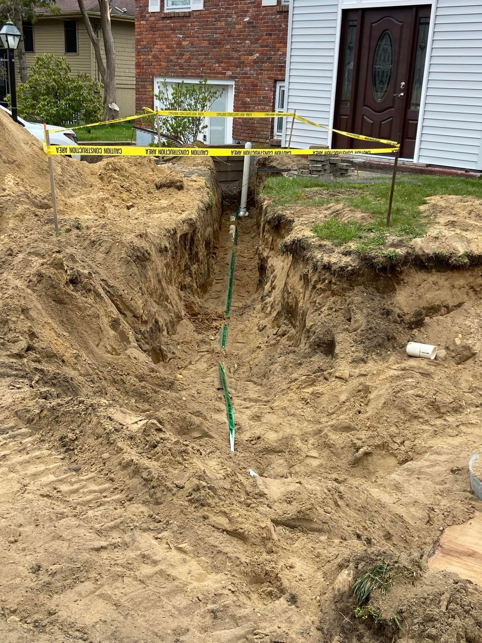 A deep trench dug in front of a house. Yellow caution tape is strung along the top. Green pipe visible in the trench.