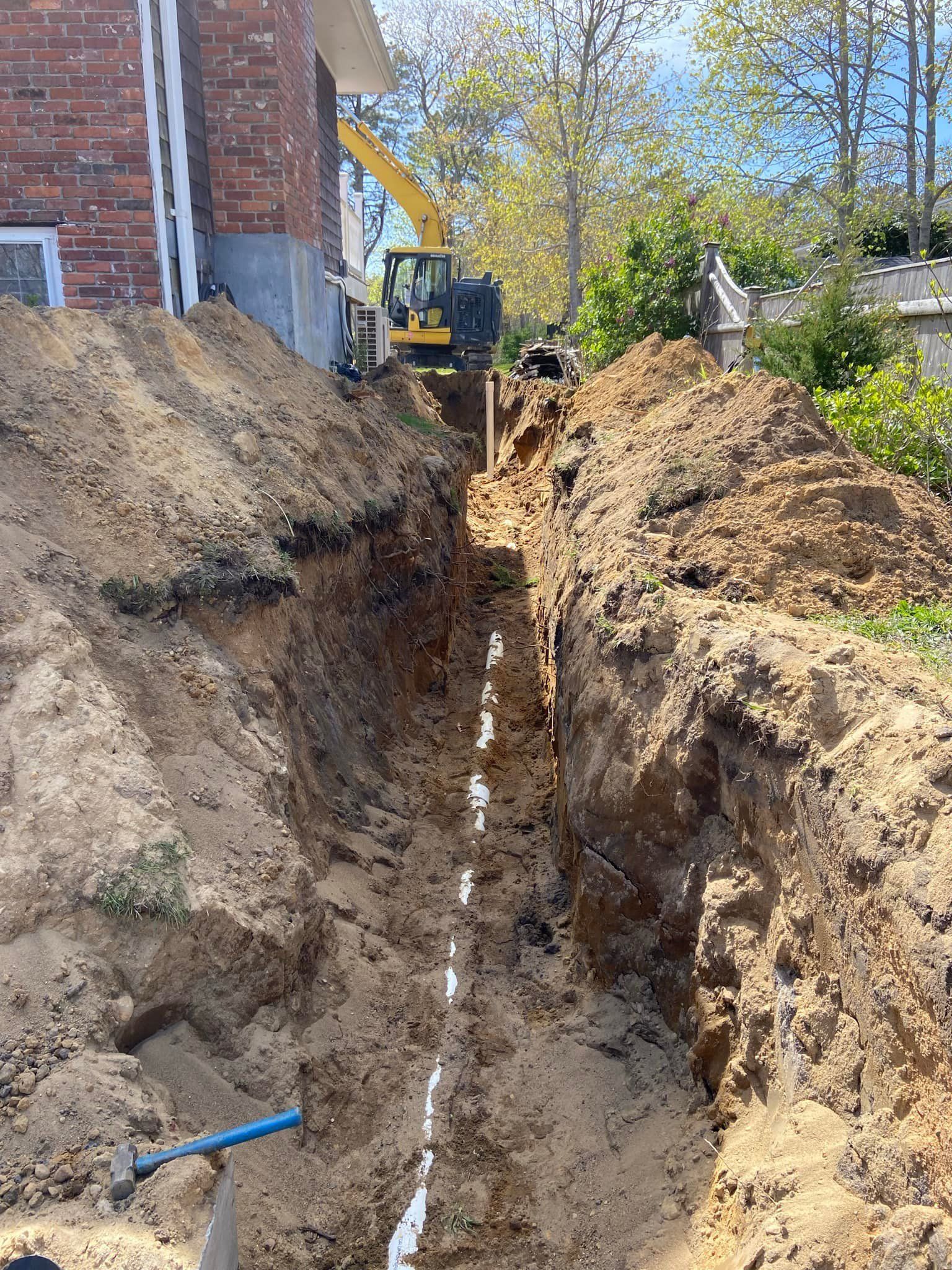 Trench dug next to a brick building for pipe installation, with an excavator in the background.