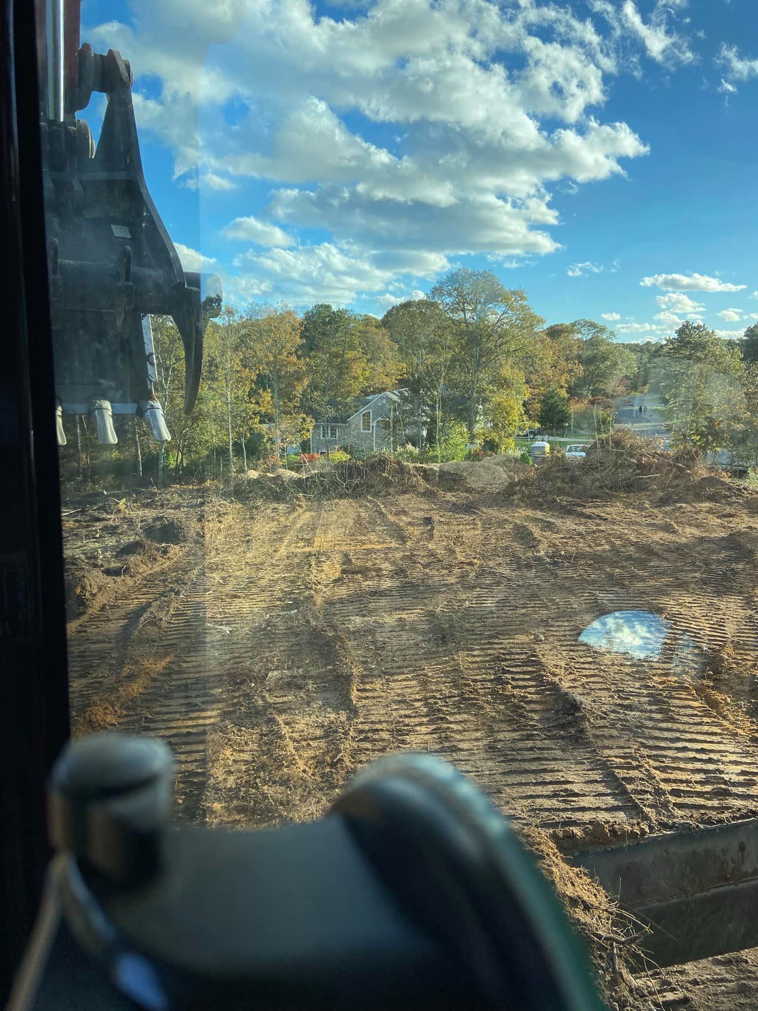View from construction equipment: dirt and trees under a cloudy blue sky.