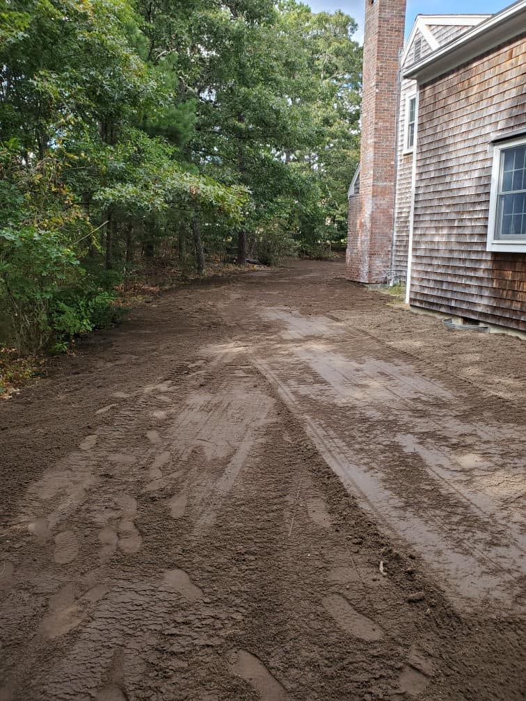 Dirt driveway with tire tracks, trees on the left, and a shingled house on the right.