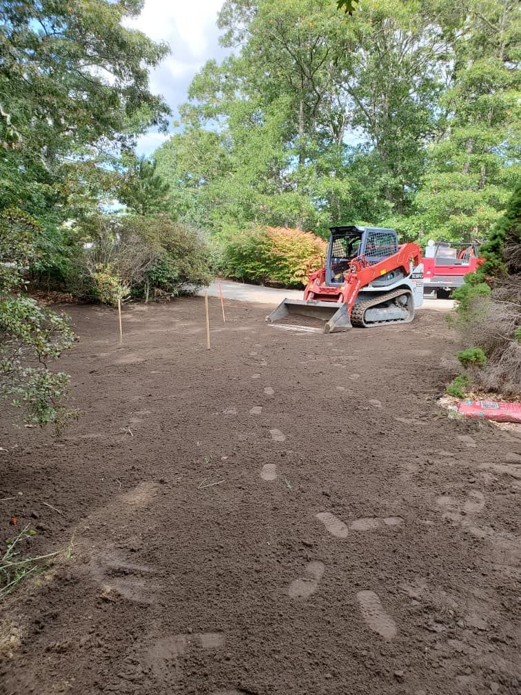 A small excavator on a leveled dirt lot, preparing a garden area. Green trees and a red truck are in the background.