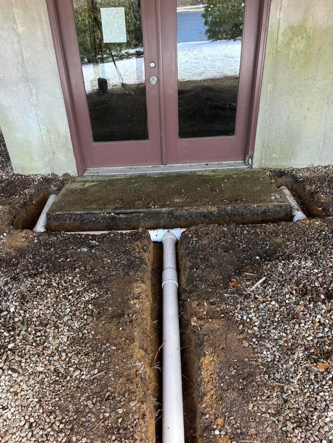 Trench with drainage pipe leading from concrete step in front of brown doors.