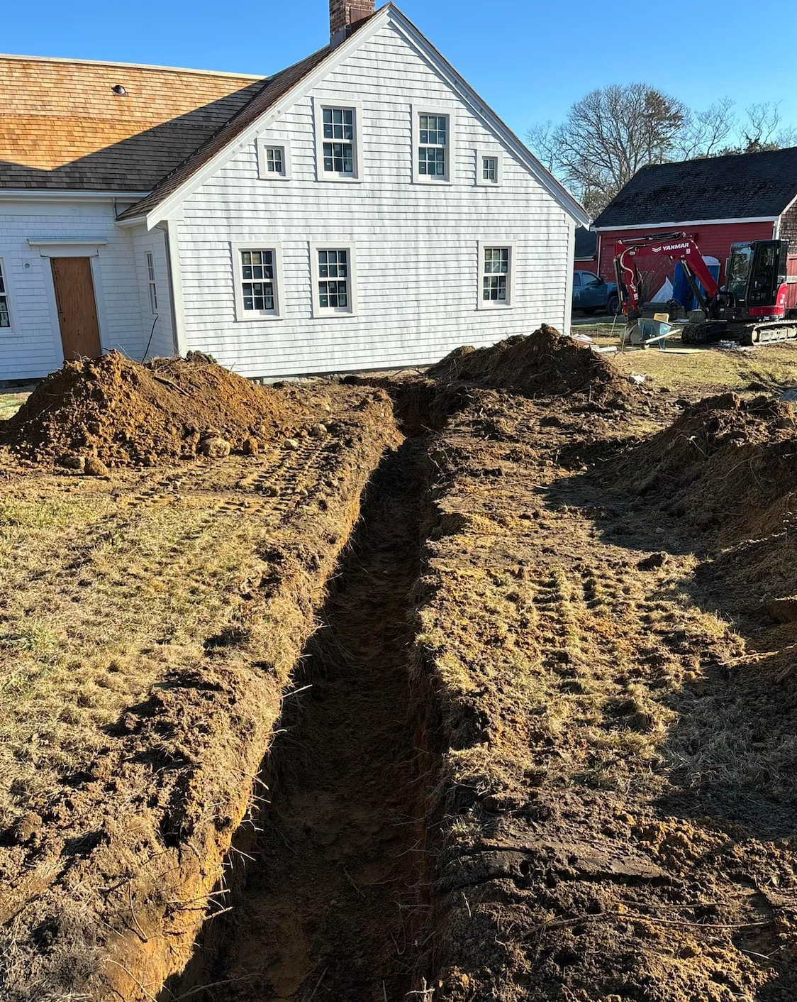 A long trench dug in a yard with a white house in the background. Piles of dirt flank the trench.