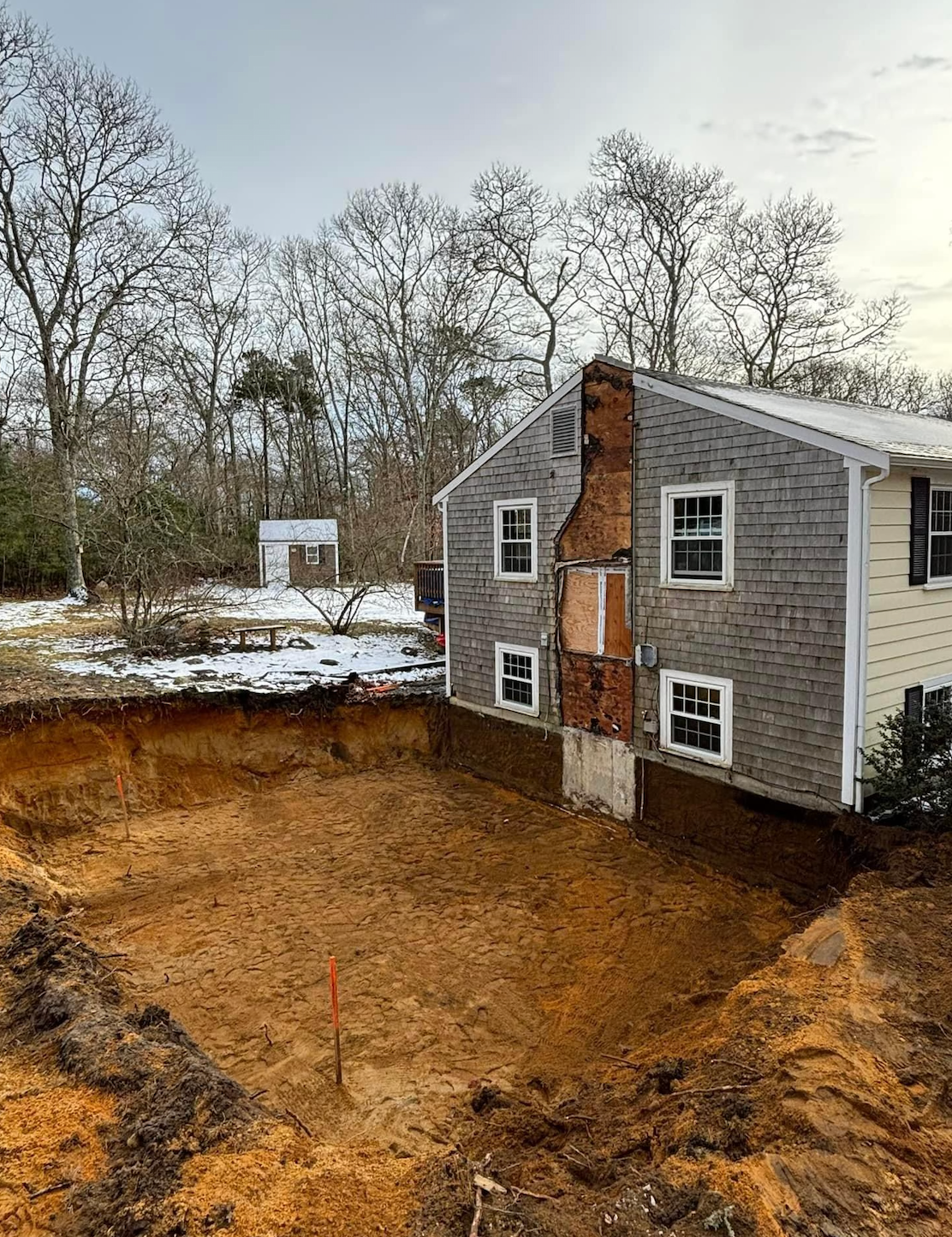 Excavation site next to a two-story house with a brick chimney; snowy ground, trees in the background.