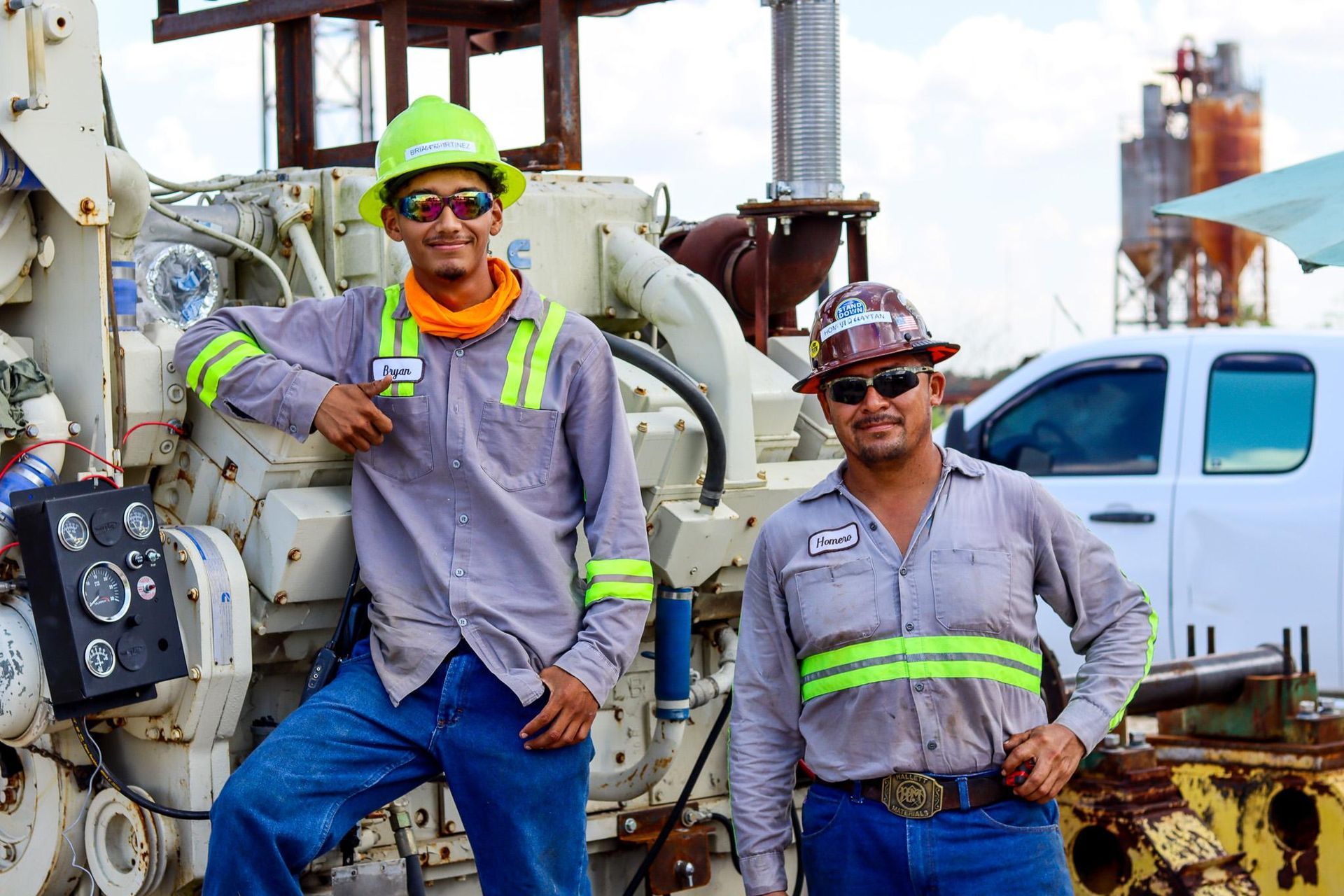 Two workers in safety gear pose by machinery at a work site. One leans; the other stands with a hand on his hip.