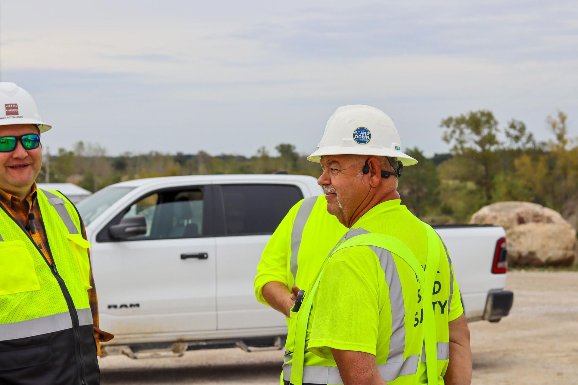 Worker in a hard hat and reflective vest stands near a vehicle on a dry, industrial site.