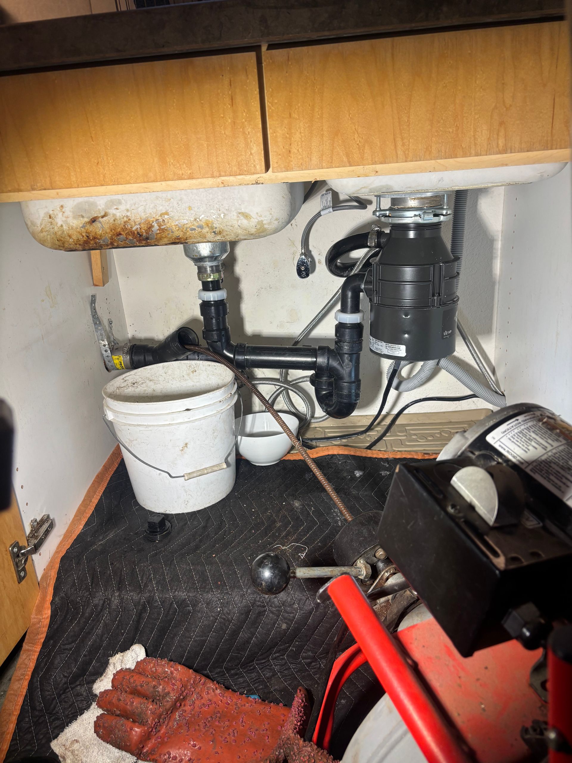 Under-sink plumbing with garbage disposal, white bucket, and red gloves on a black mat