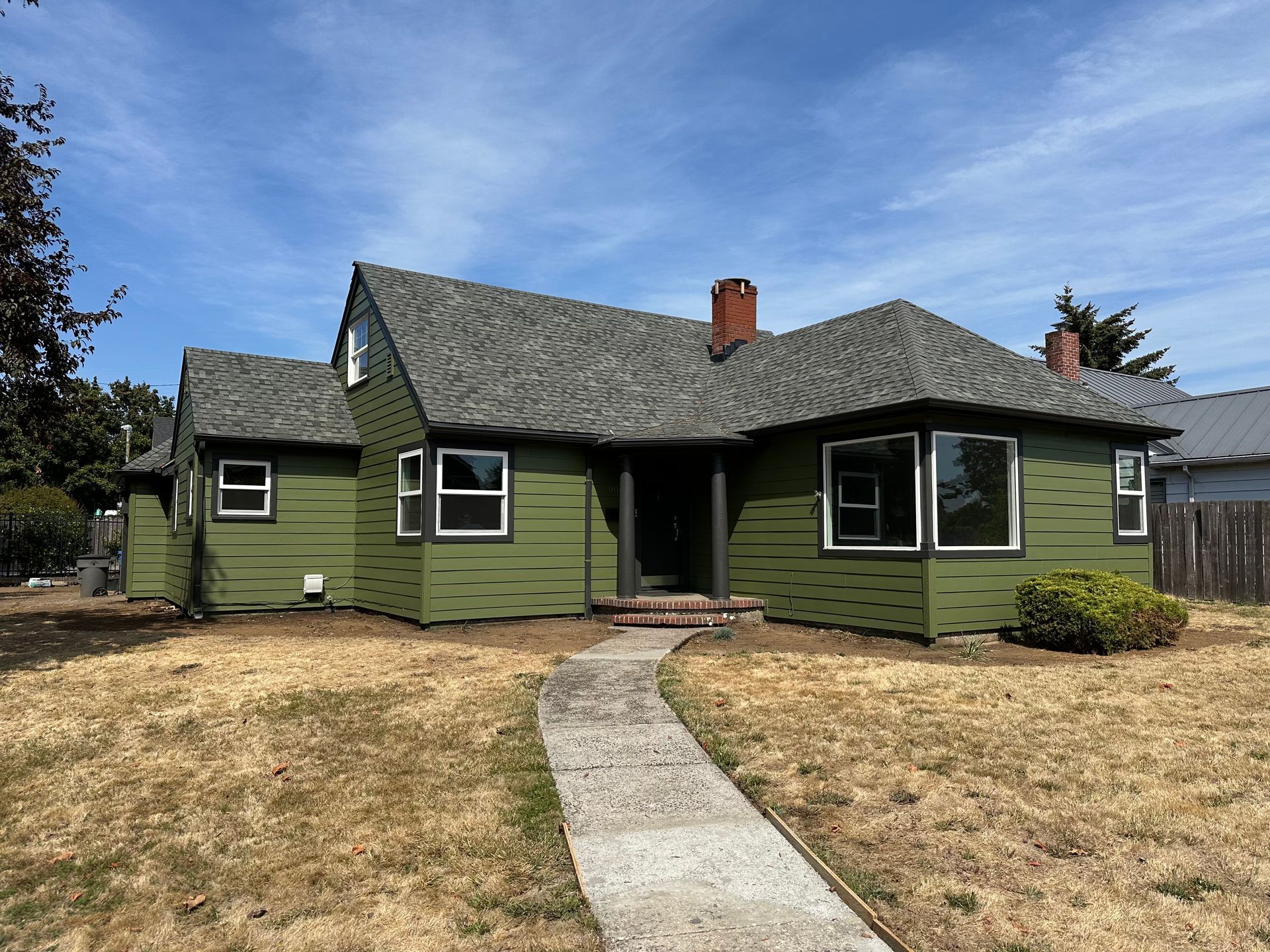 Green single-story house with a gray shingled roof and a curved concrete walkway under a blue sky