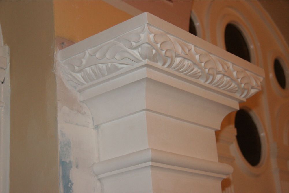 Decorative white column capital with ornate carvings against an orange wall and arched windows