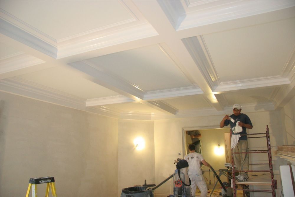 Workers painting a cream-colored room with ornate ceiling moldings and a ladder in use