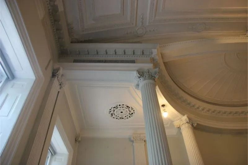 Ornate white interior ceiling with a tall column, decorative molding, and a large window on the left