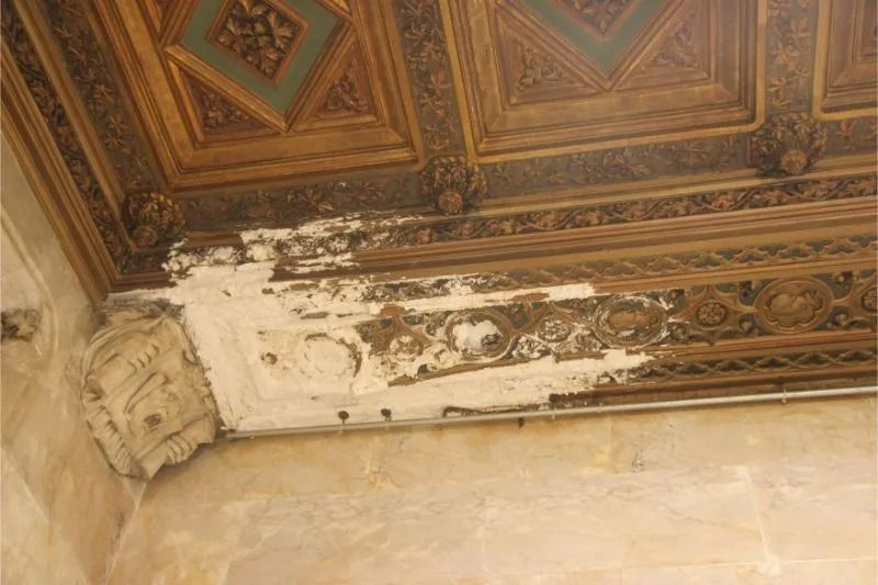 Ornate ceiling corner with peeling paint and decorative woodwork above a beige wall
