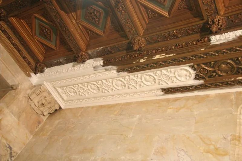 Ornate carved wooden ceiling with white decorative molding above a beige marble wall