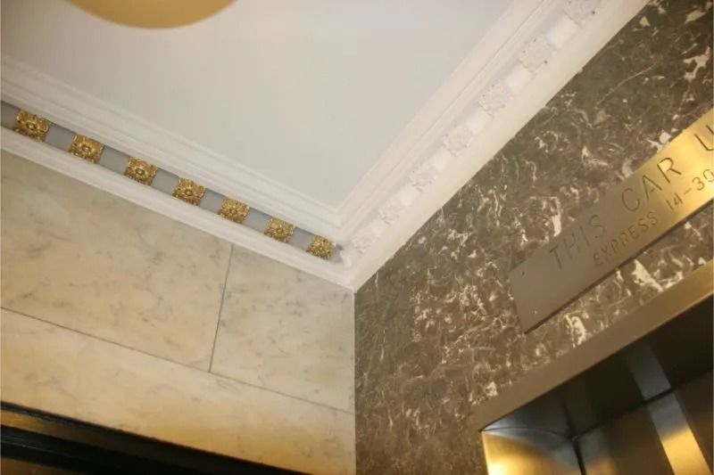 Interior corner with ornate gold trim, marble wall, and a metallic plaque above an elevator door