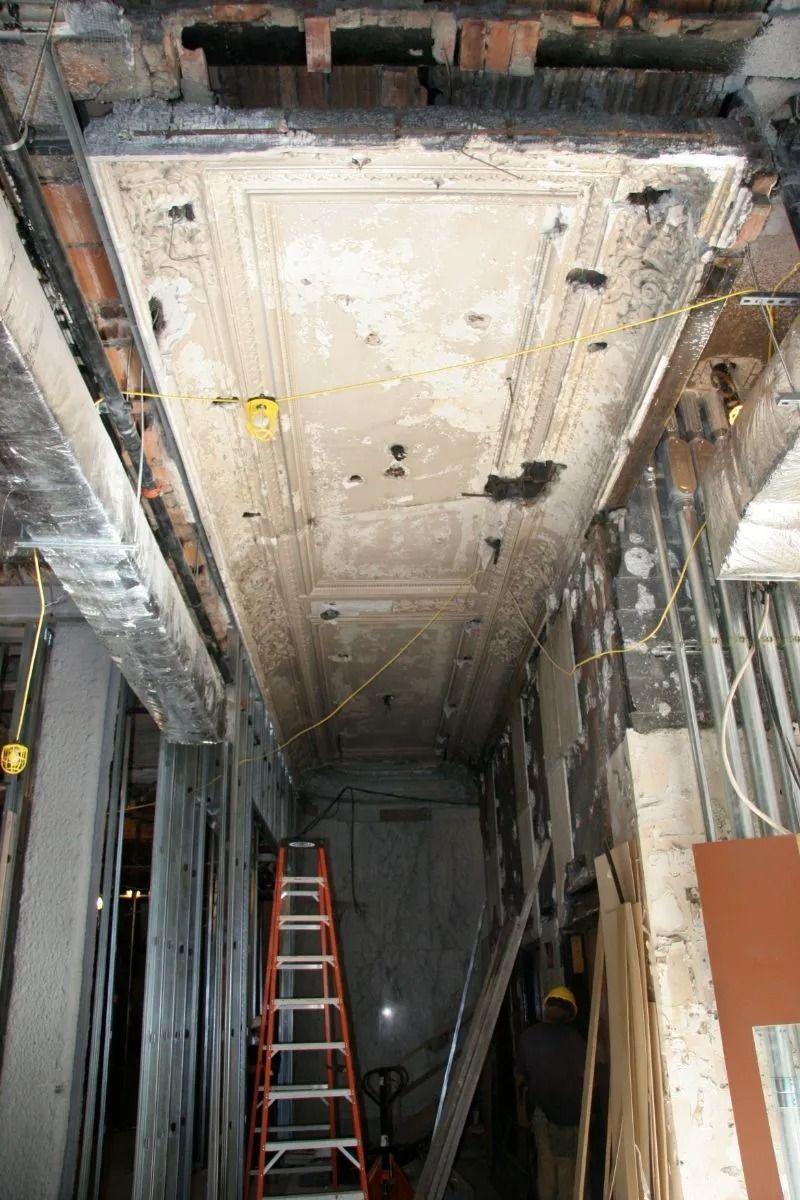 Damaged ceiling panel in a narrow hallway, with a red ladder below and exposed framing above