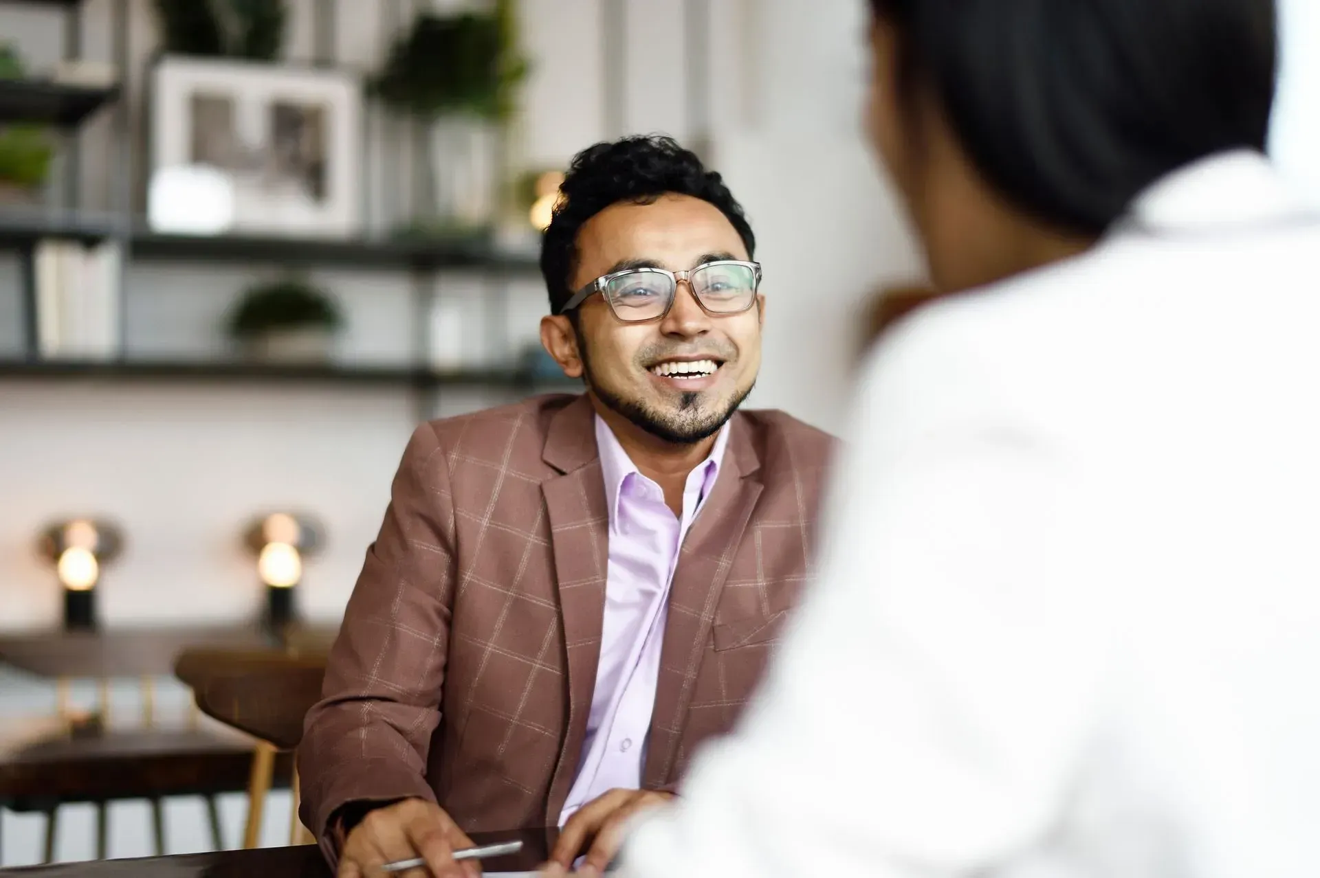 Man in brown blazer smiles while speaking to a person in a white blazer at a table. Man in brown blazer smiles while speaking to a person in a white blazer at a table.