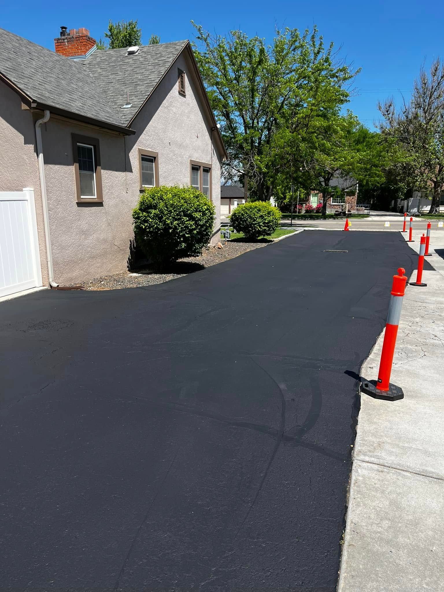 A newly paved black asphalt driveway next to a light-colored stucco house, marked by several orange traffic cones.