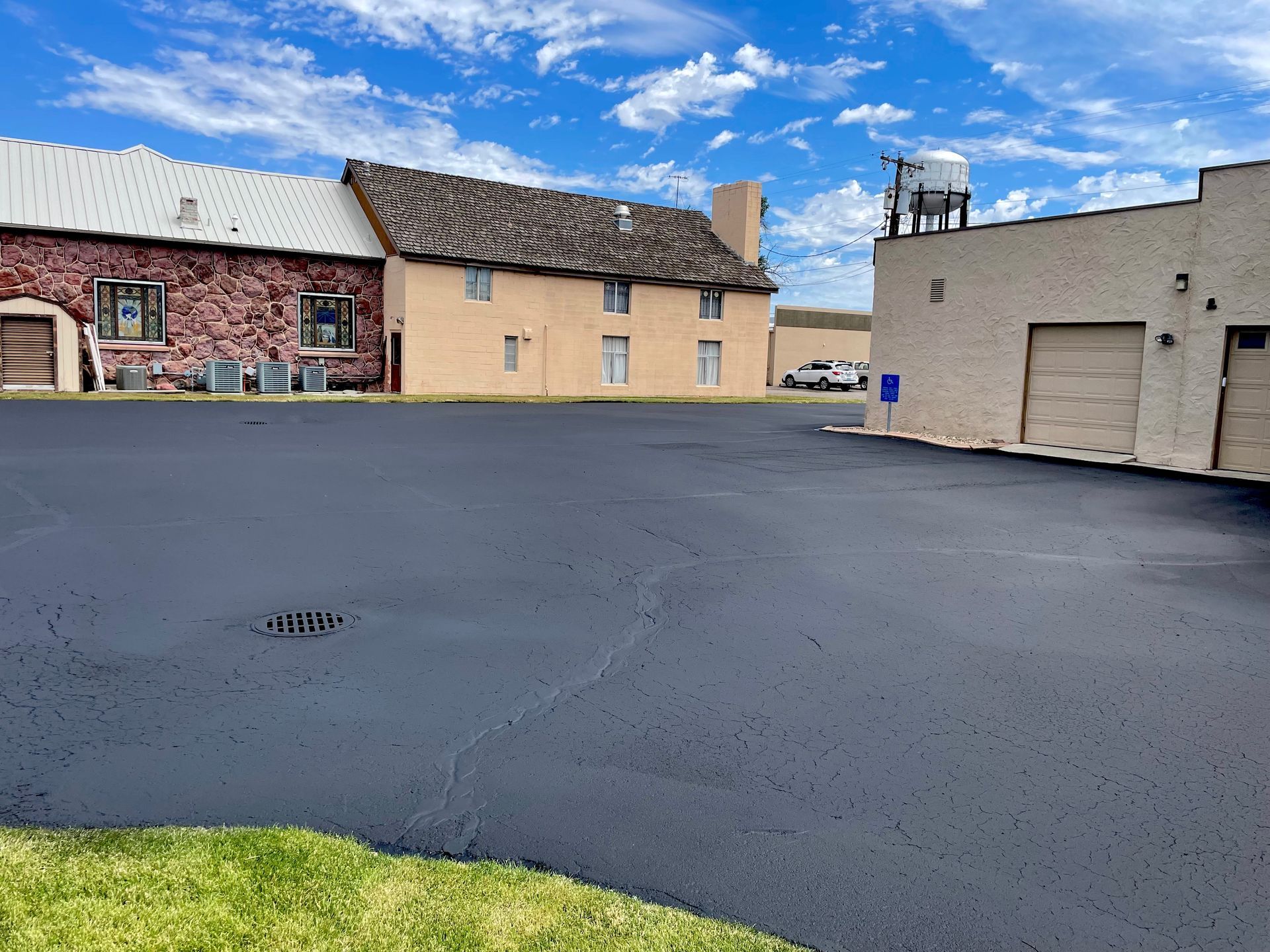 A freshly paved black asphalt parking lot in front of two historic-looking buildings under a bright blue, cloudy sky.