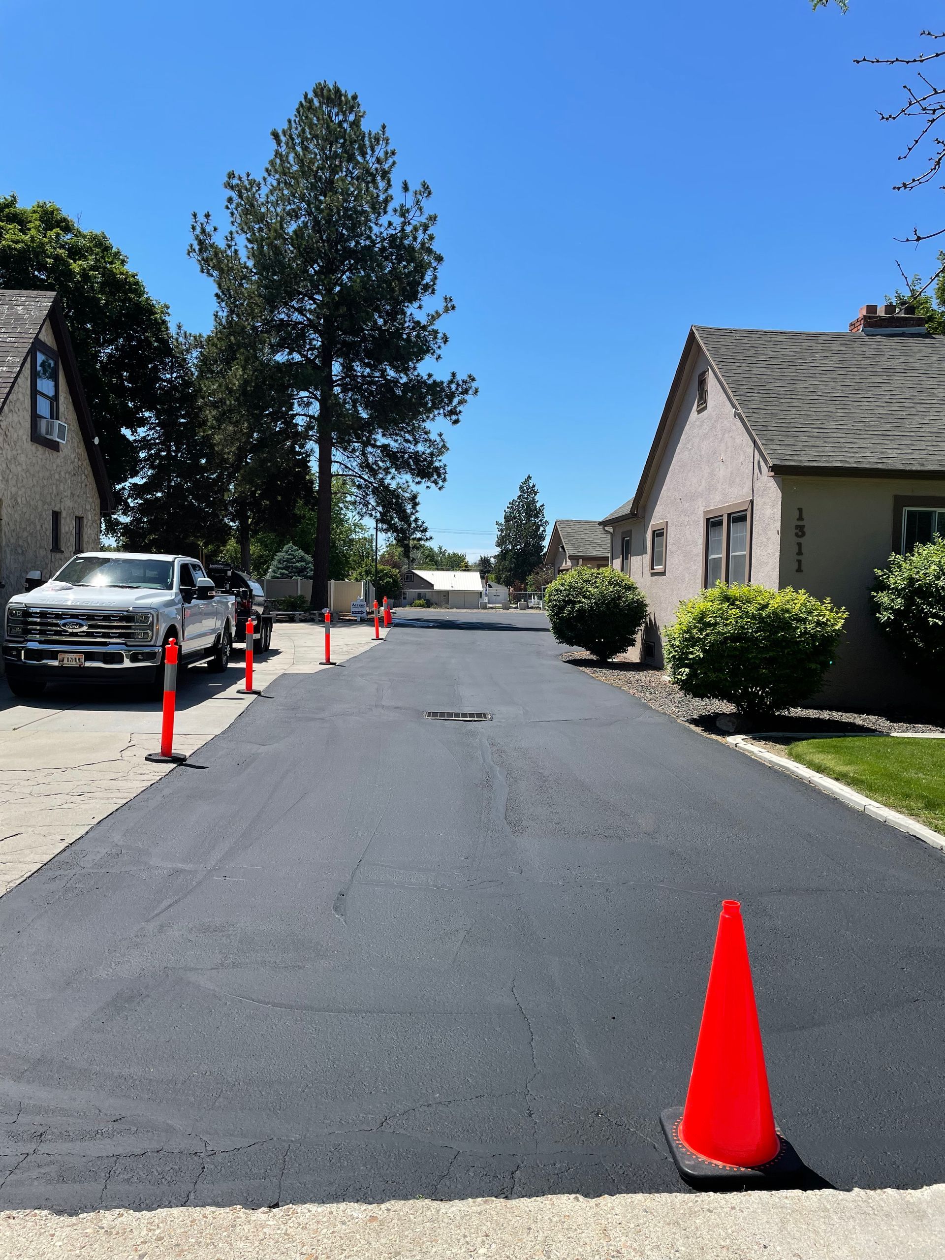 A newly paved asphalt driveway between two houses, marked with orange traffic cones during the day.