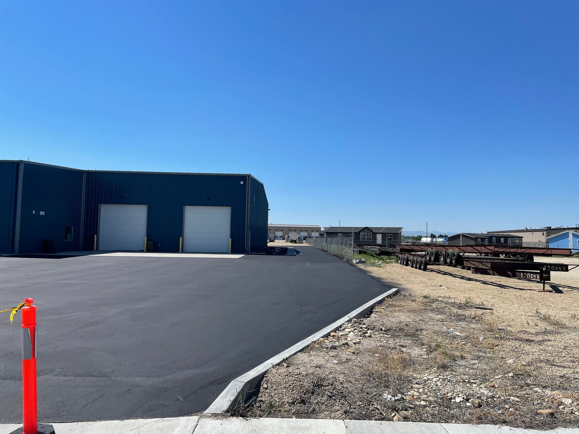 A blue warehouse with two white doors next to a newly paved asphalt parking lot under a clear blue sky.